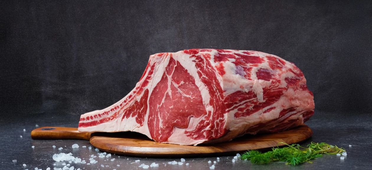A raw ribeye steak resting on a wooden cutting board with herbs