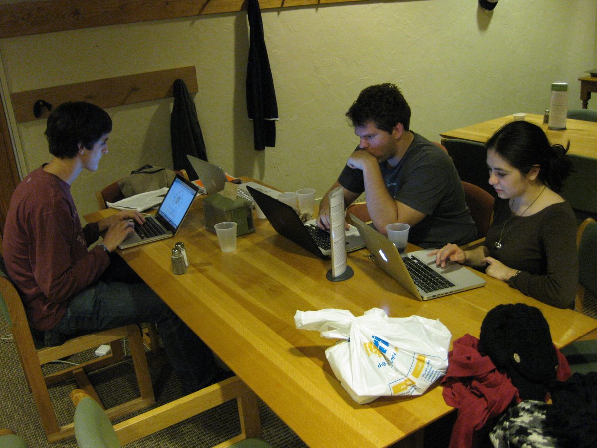 Three students sit at a table with laptops, each working alone