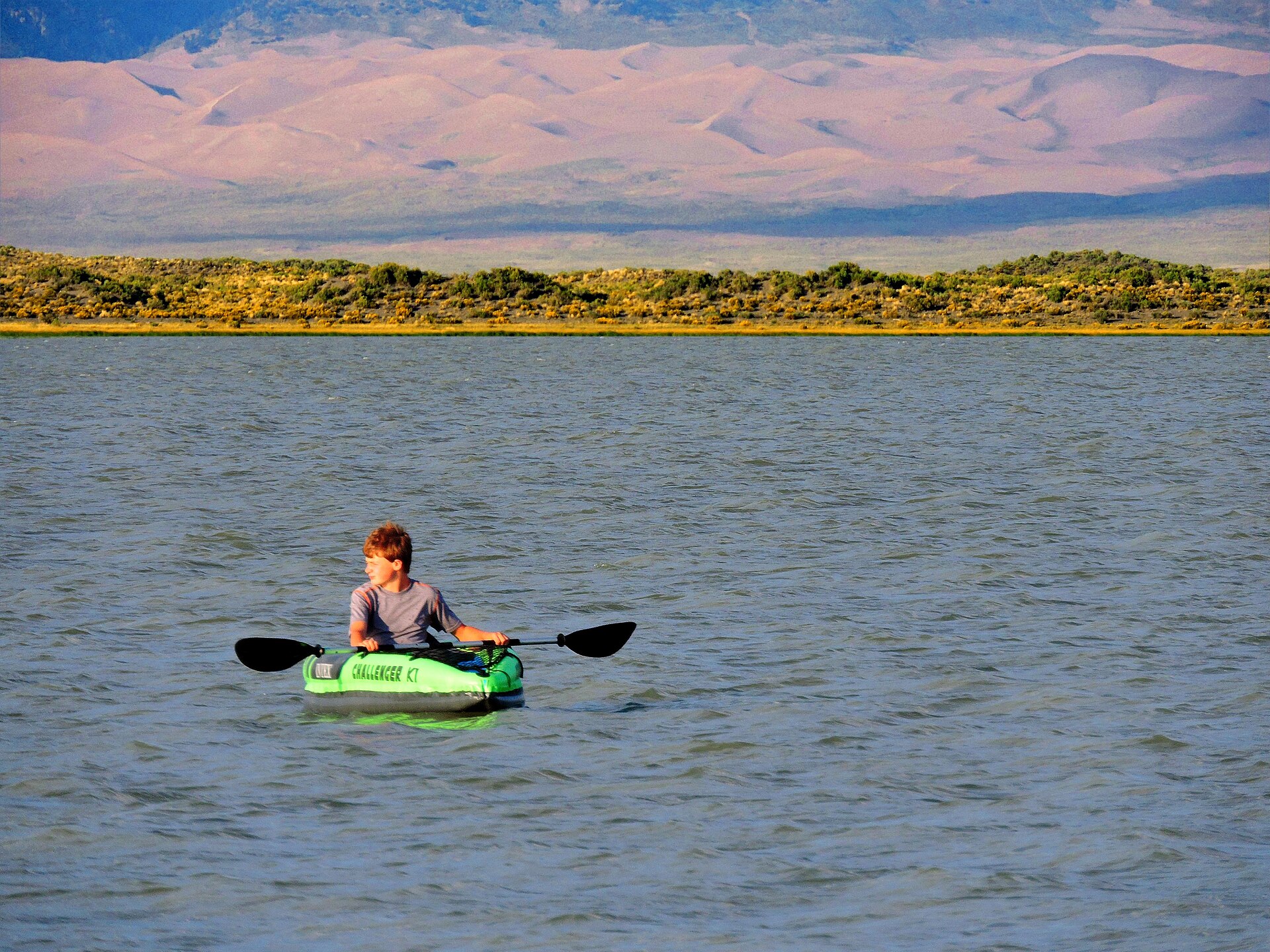 A kayaker paddling on calm water with dunes in the background