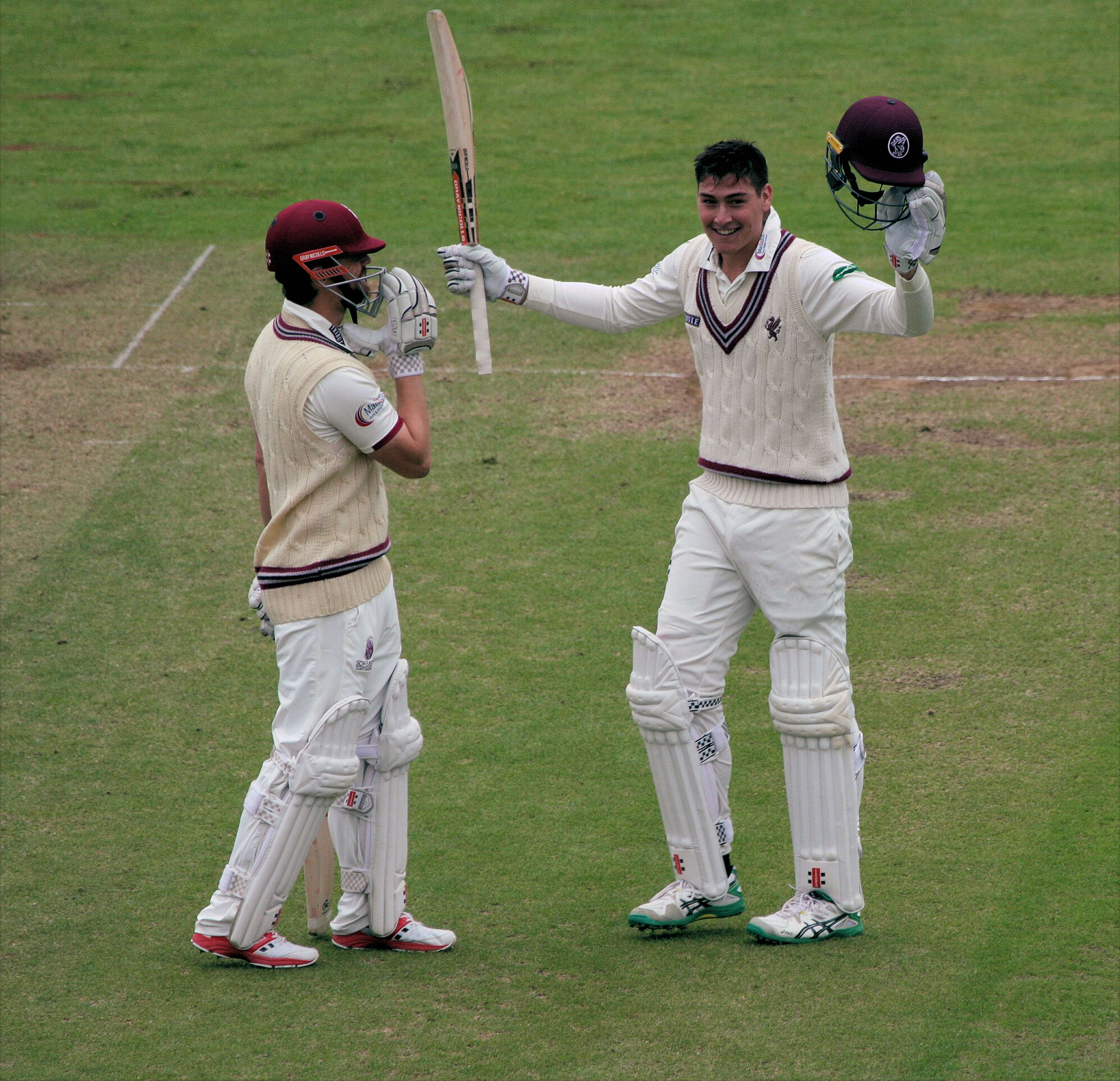 A cricket batter celebrates reaching a century with bat and helmet raised