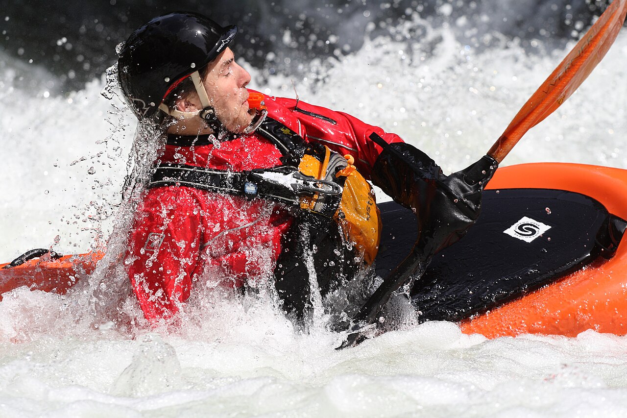 A kayaker mid-roll in whitewater, water splashing off his helmet and life vest