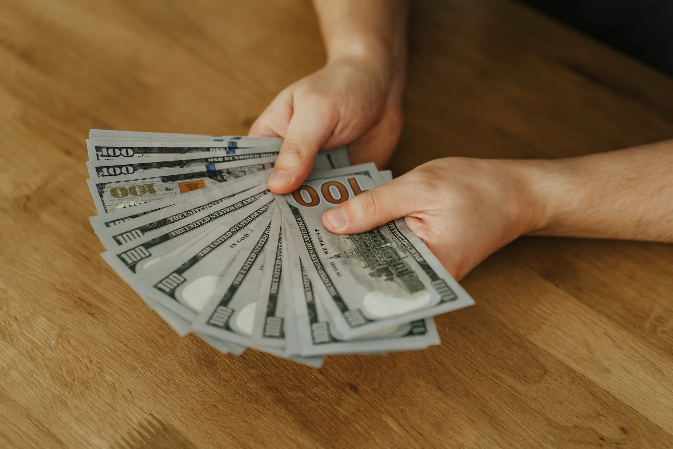 Hands spreading a fan of one-hundred-dollar bills on a wooden table
