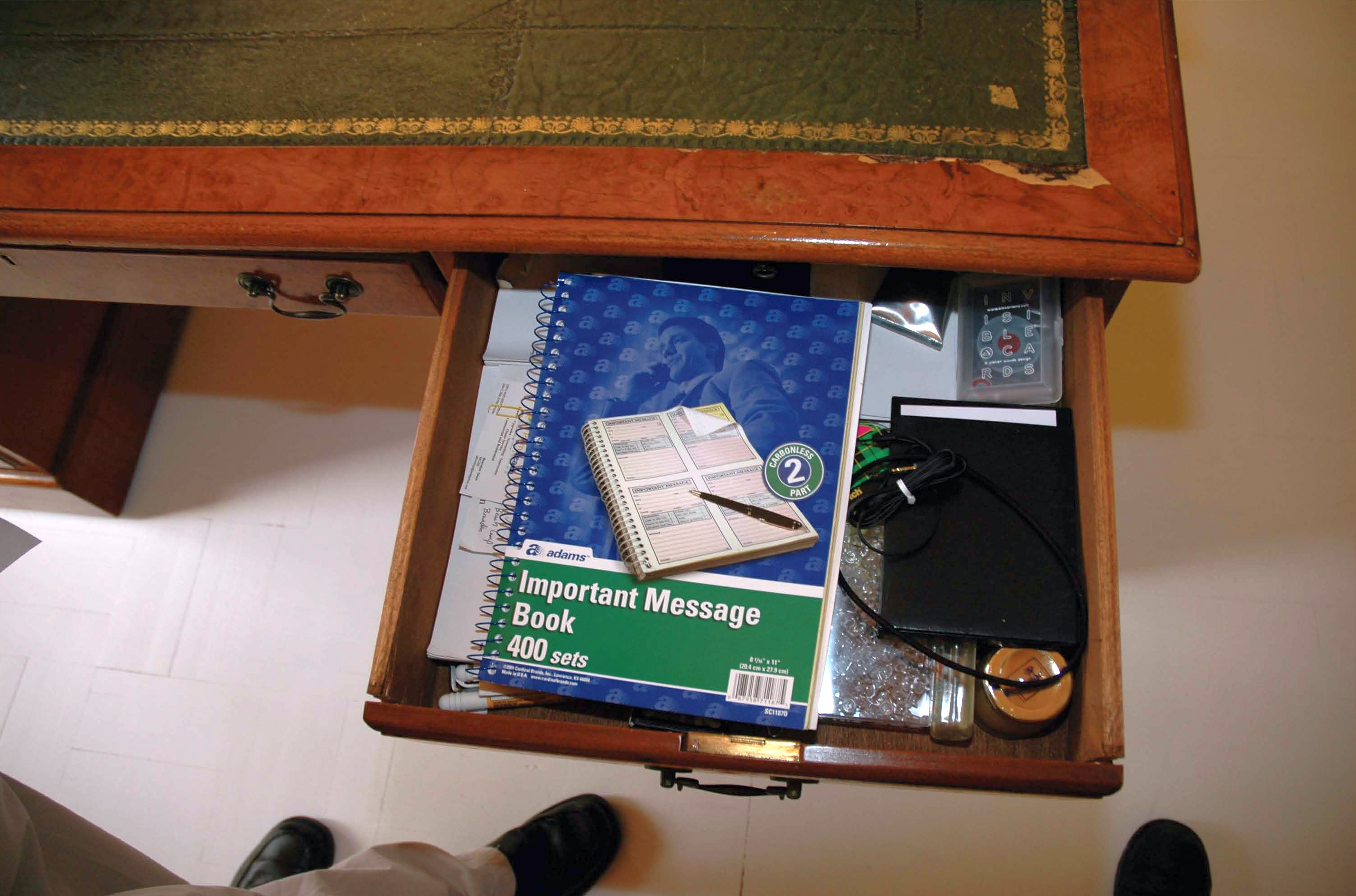 An open wooden desk drawer with notebooks, stacked papers, and a pen