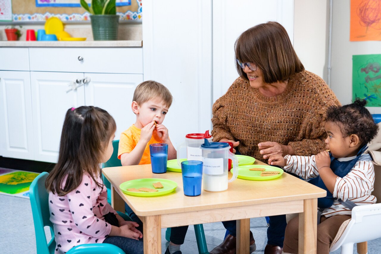 A childcare provider sits at a snack table with three young children eating crackers