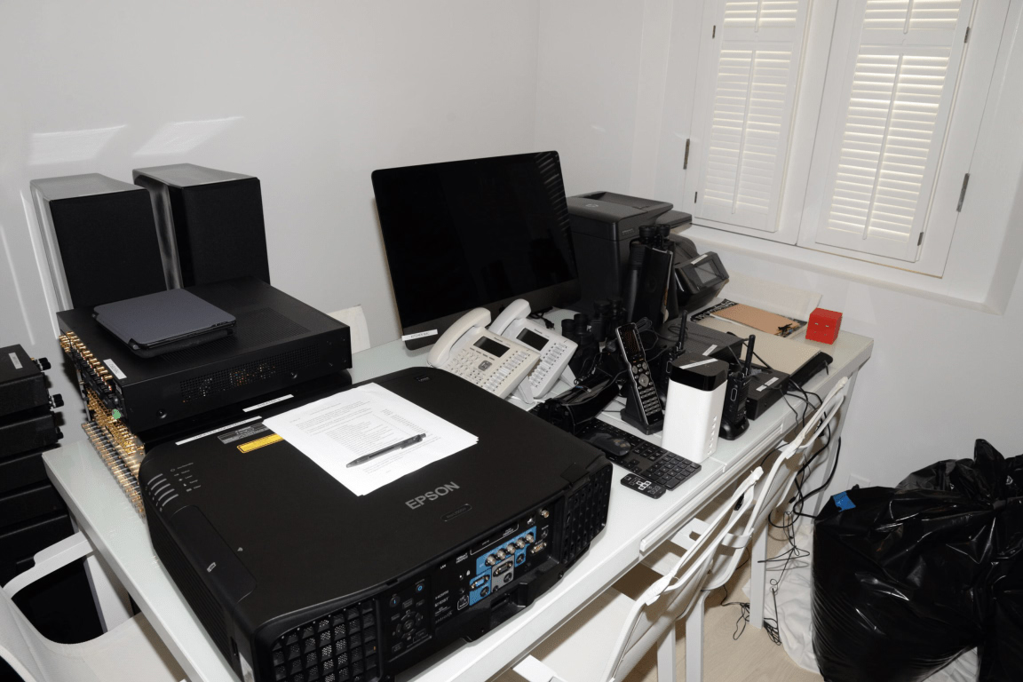 A desk with a projector, monitor, and devices crowded together beside a window