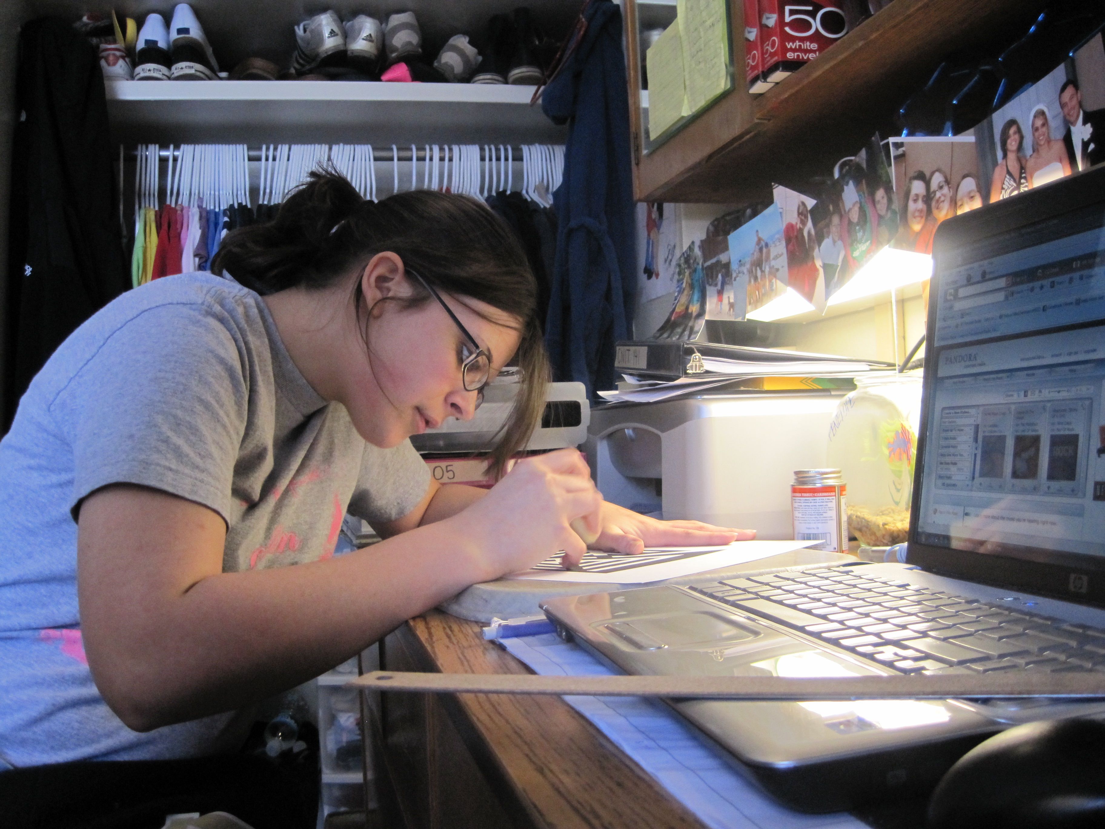 A student writing at a desk in a dorm room, studying with books and notes spread out