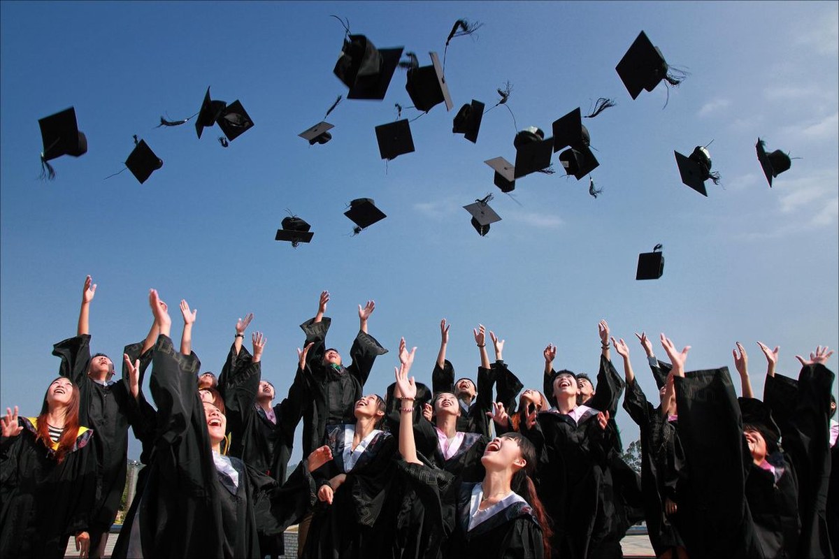 Graduates in black gowns throw their caps in the air on a sunny day.