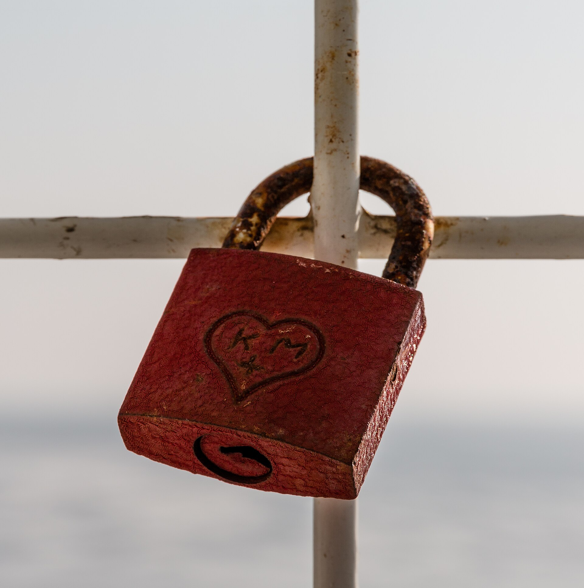 A weathered red padlock with a heart engraving hanging on a white railing