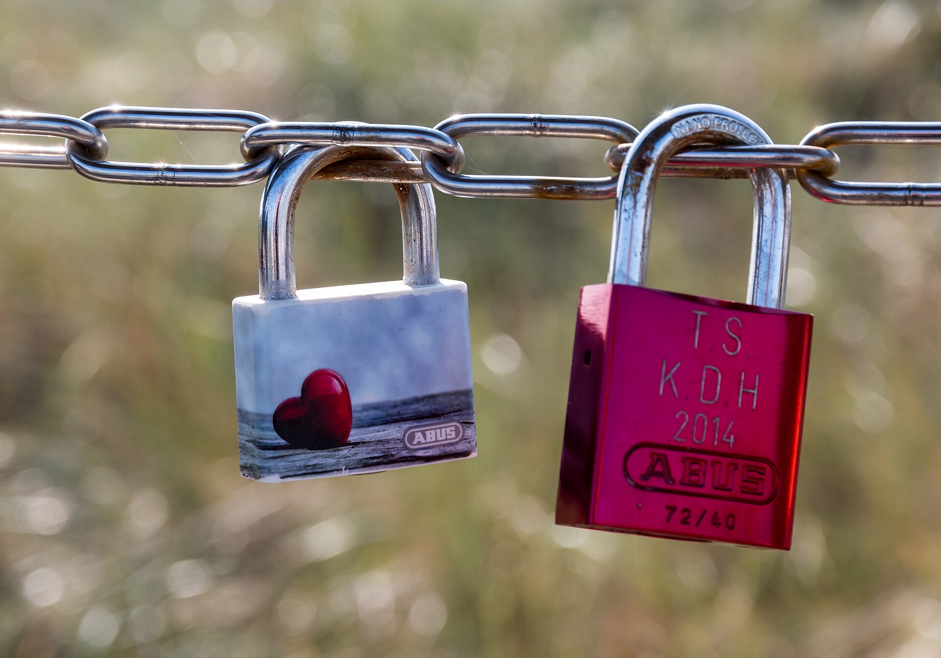 Two padlocks on a chain, one white with a red heart and one pink with engraved letters