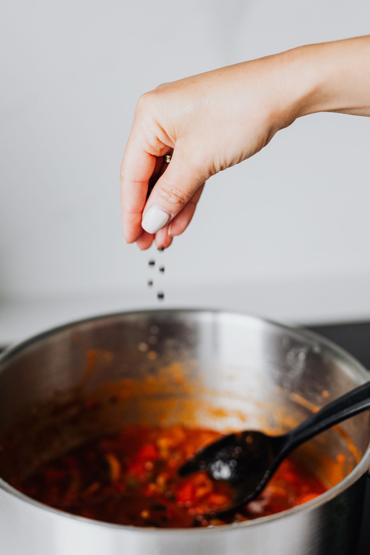 A hand seasons tomato soup with black pepper in a stainless steel pot