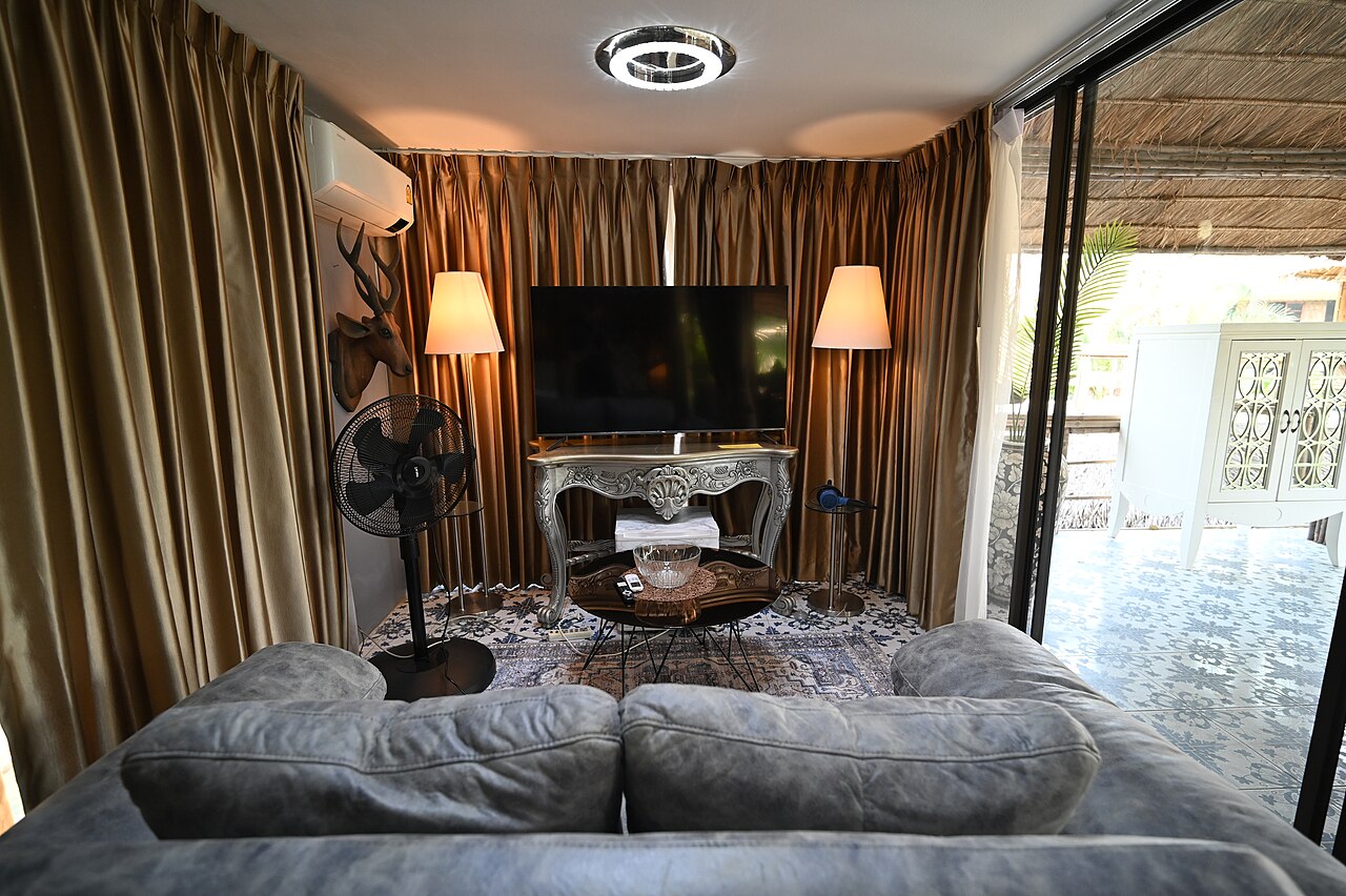 DSC 8584 Cozy living room with plush gray sofa facing a TV on an ornate console warm floor lamps and floor-to-ceiling curtains opening to a sunlit patio