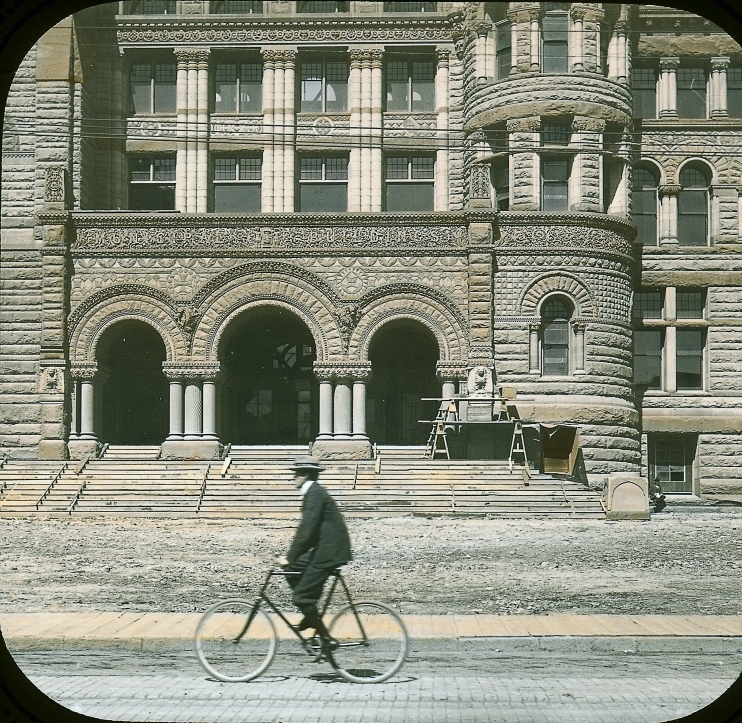 A cyclist riding past a grand stone building on a city street in a historic photograph