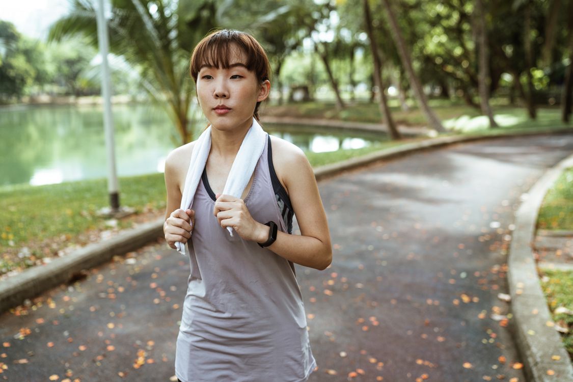 An athletic woman jogging outdoors in a park with a towel around her neck