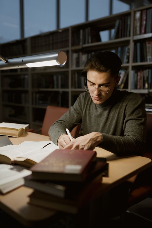 A young man studying in a library with books and a desk lamp