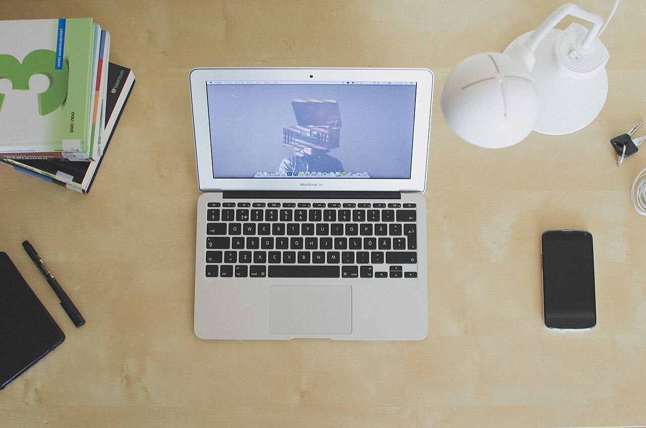 A laptop on a desk beside a lamp, books, and a phone