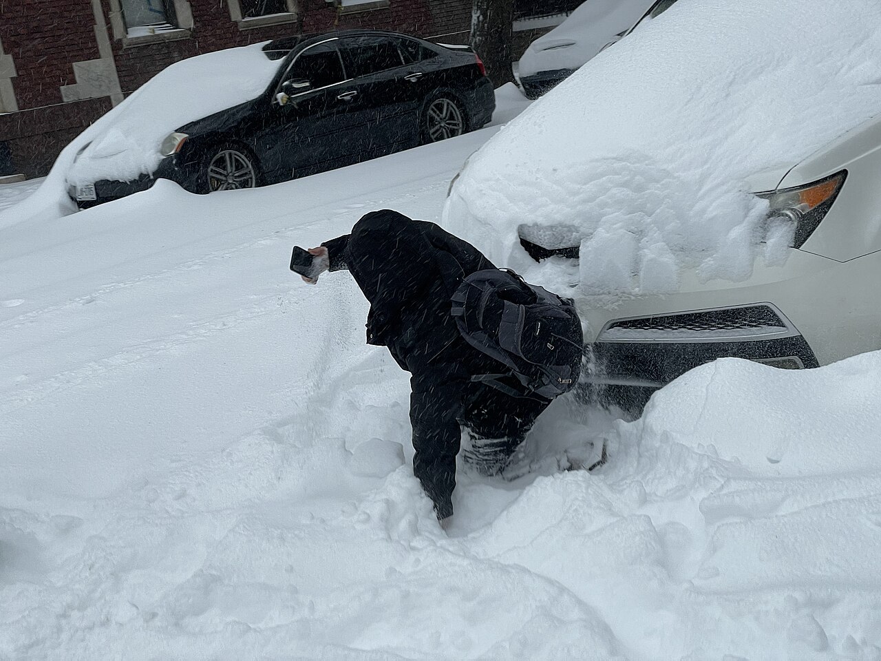 A person holding a smartphone in deep snow beside snow-covered cars