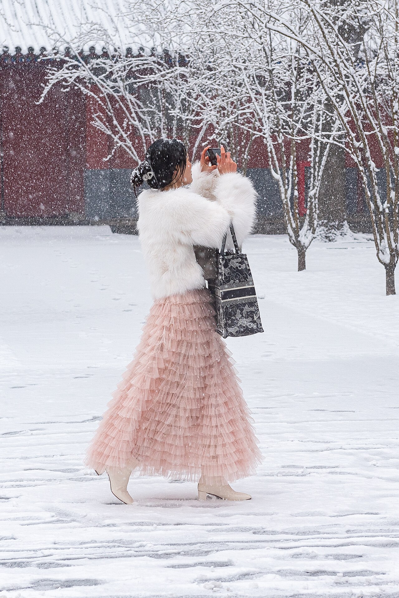 A woman photographs snow-covered trees with her phone during snowfall
