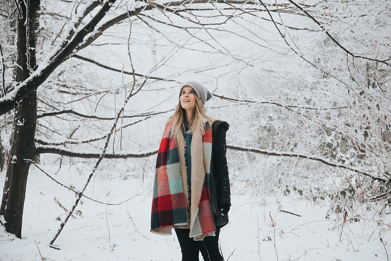 A woman in a knit hat and a large plaid scarf standing among snow-covered trees