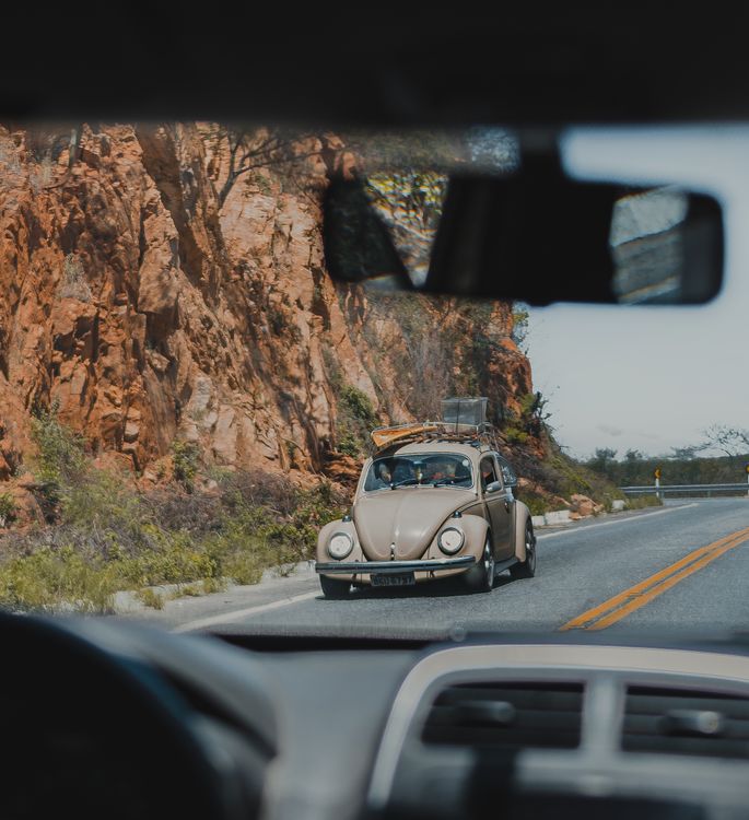 A classic car on a road beside rocky cliffs, seen through a windshield