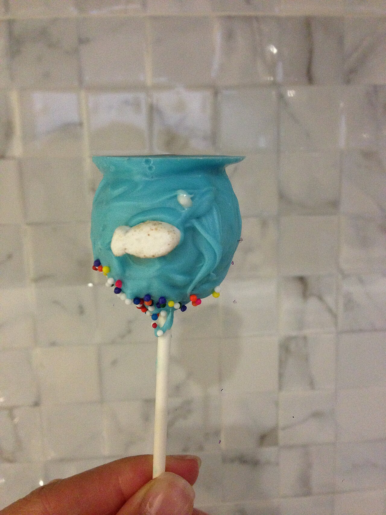 A blue fishbowl cake pop with sprinkles held up in a kitchen