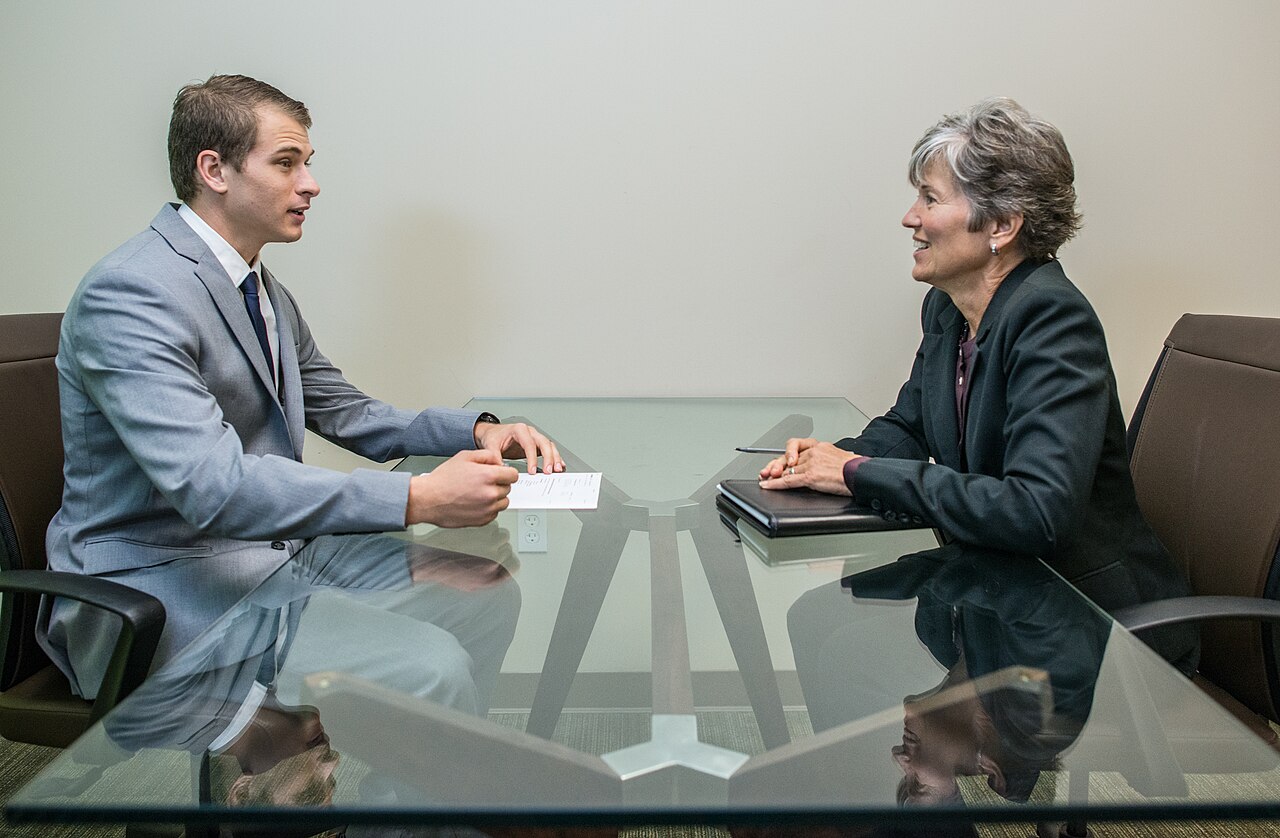 A man in a gray suit speaking with a woman across a glass table during a job interview
