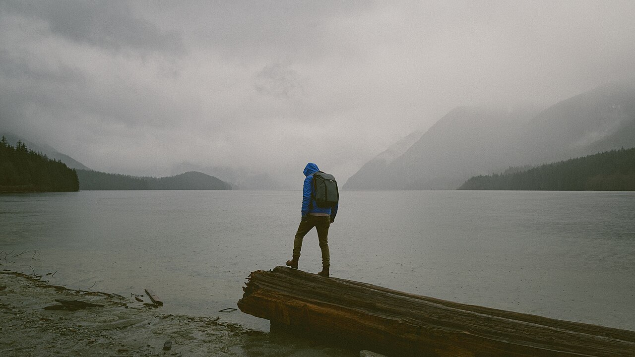 A hiker stands on a log near a lake in mist and light rain