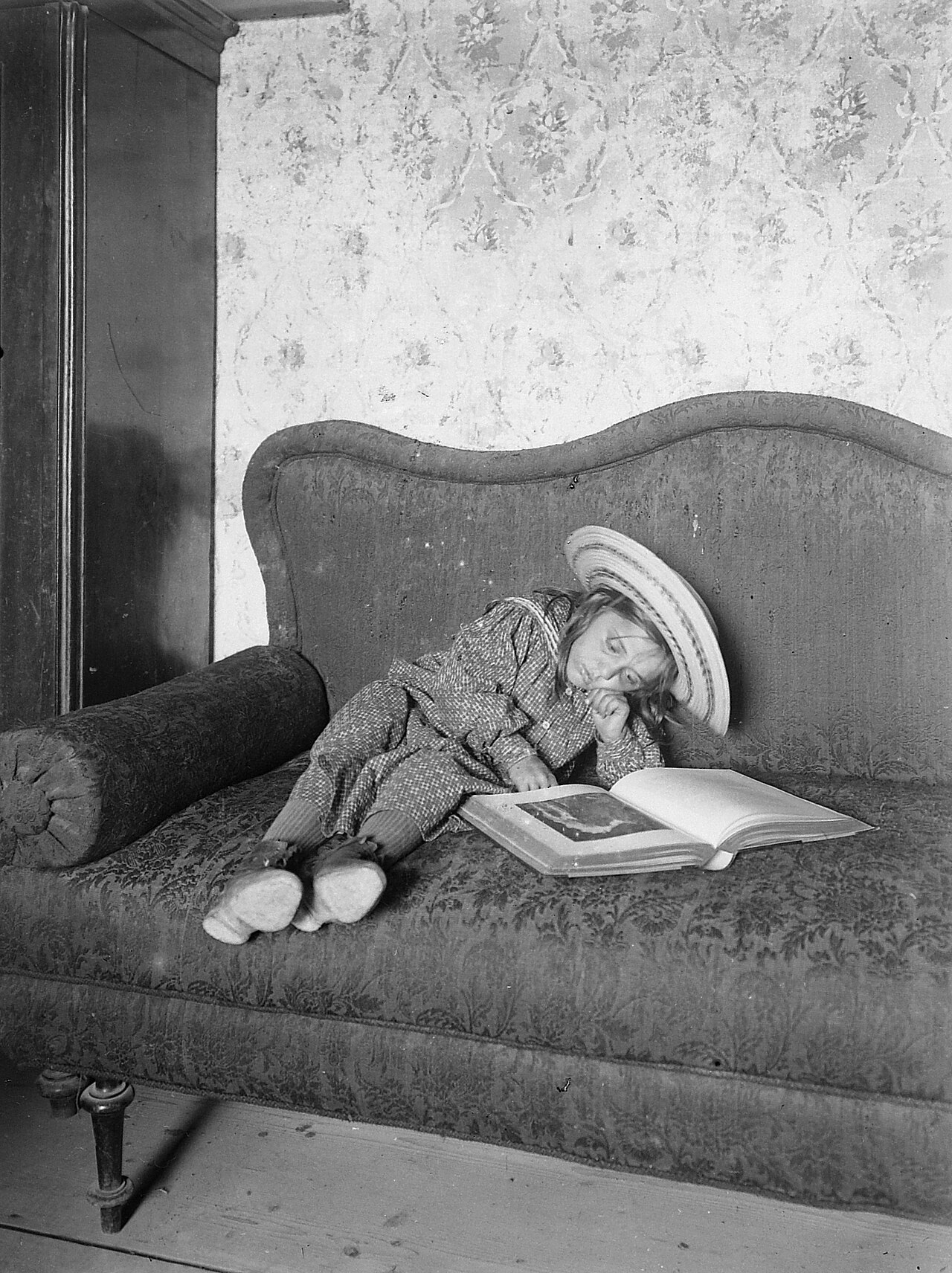 A child in a hat reading a large book on a sofa
